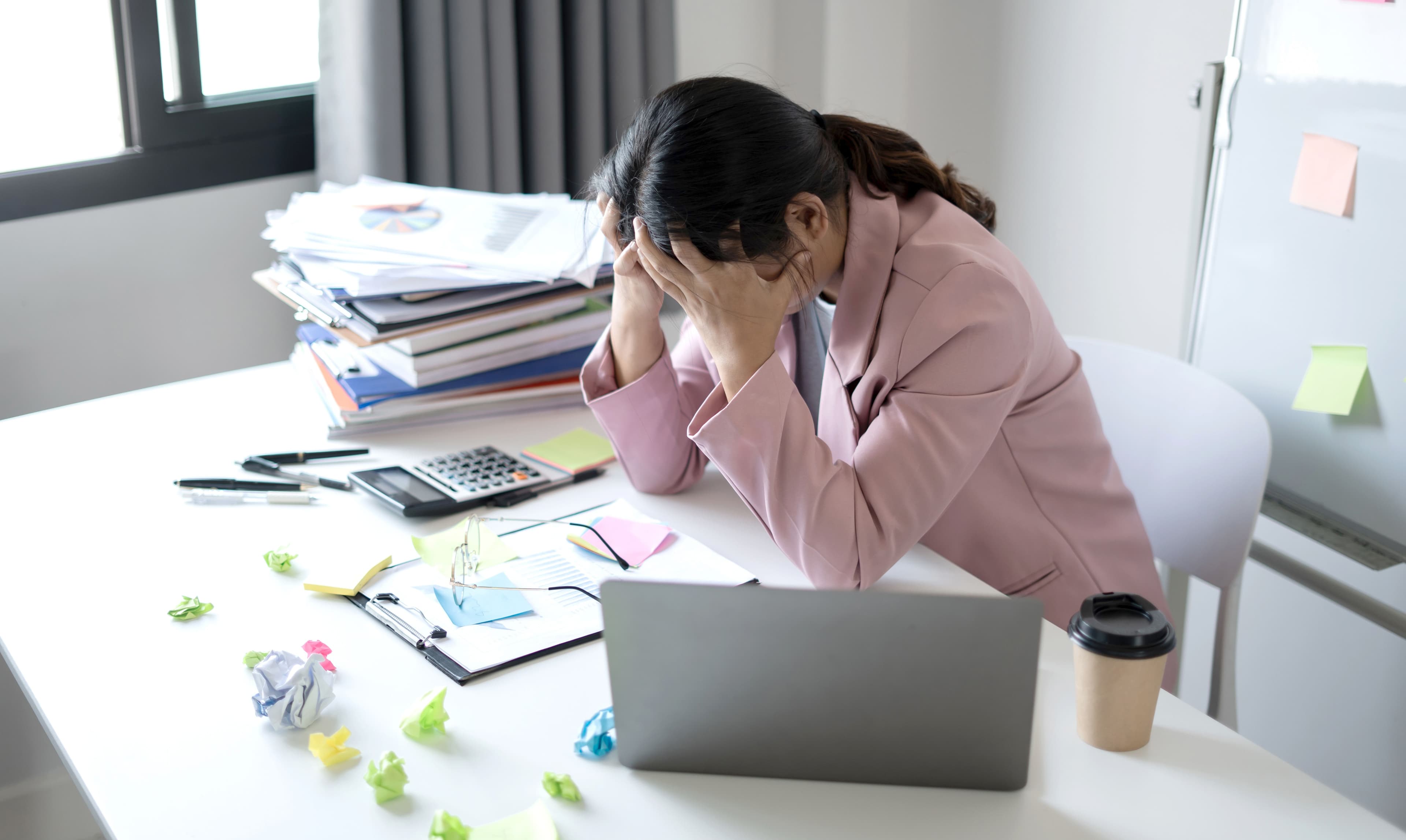Female office worker sitting by her table holding both hands on her head in distress.